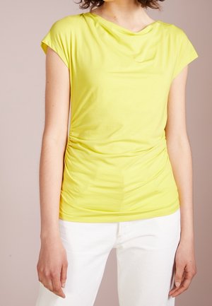 Woman wearing a bright yellow short-sleeve top with side ruching paired with white pants against a plain beige background.