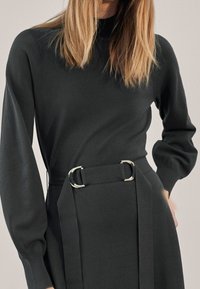 Robe en tricot gris foncé avec un col montant, des manches longues ajustées et une ceinture avec des éléments en métal argenté circulaires à la taille.