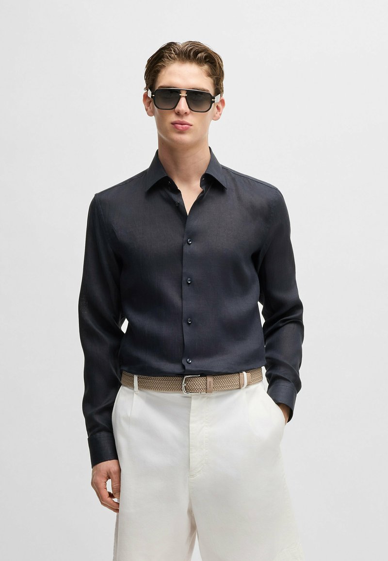 Camicia blu scuro a maniche lunghe con chiusura a bottoni, tessuto texturizzato, abbinata a pantaloni bianchi e una cintura di paglia beige. Occhiali da sole neri indossati.