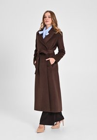 Cappotto lungo in misto lana marrone con cintura, colletto scialle e tasche laterali, indossato sopra pantaloni neri e tacchi beige.