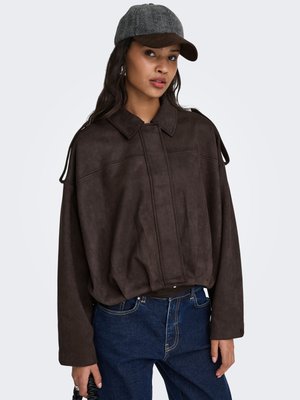 Jonge vrouw met een grijze visgraatpet, donkerbruine oversized jas en blauwe spijkerbroek, poserend tegen een effen lichte achtergrond.