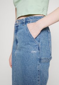 Osoba nosící světle zelený krátký top a modrou džínovou sukni s rukou v kapse, na přední straně sukně vyšívané logo „obey“.