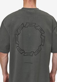Grå oversized T-shirt lavet af bomuld, med et stort cirkulært sort logo grafisk tryk på ryggen og et lille mærke nær sømmen.