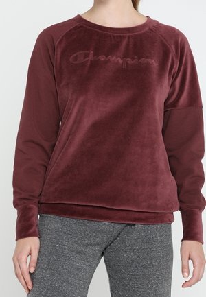 Vrouw draagt een bordeauxrode velours Champion-trui en grijze joggingbroek, staat met armen ontspannen langs het lichaam.