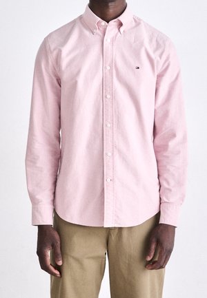 Mężczyzna w jasnoróżowej koszuli z długim rękawem zapinanej na guziki z małym logo na piersi i beżowych spodniach, stojący na jednolitym białym tle.