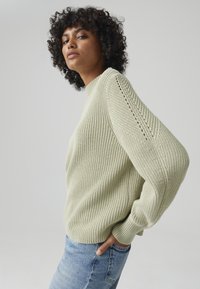 Hellgrüner Strickpullover mit hohem Kragen, strukturiertem Muster und überschnittenen Schultern. Kombiniert mit blauen Jeans, die eine lockere Passform zeigen.