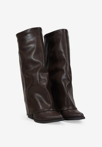 Botas de cuero de color marrón oscuro hasta la rodilla, con una textura ligeramente arrugada y punteras afiladas, de pie en una superficie blanca.