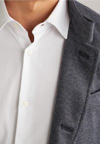 Camicia bianca con chiusura a bottoni sotto un blazer blu scuro testurizzato con motivo a spina di pesce e revers a punta.