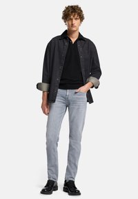 Chemise en denim foncé avec les manches retroussées sur un polo noir, associée à un jean bleu clair et des chaussures à lacets noires, se tenant devant un fond blanc.