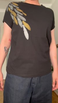 Camiseta negra de manga corta con un diseño de pluma en gris, oro y blanco en el hombro izquierdo. Tejido suave con un corte relajado.