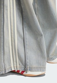 Light blue denim pants with white side stripes, featuring a zipper detail at the hem. Accented with red stripes on white shoes.