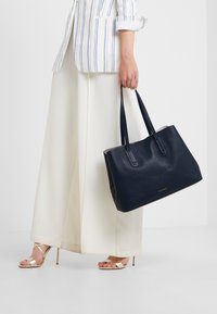 Granatowa skórzana torba tote z dwoma uchwytami u góry, o fakturowanej powierzchni i minimalistycznym logo na dole. Noszona z białym strojem.