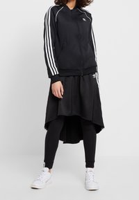 Svart zip-up jacka med vita randiga ärmar, matchad med en flödande svart asymmetrisk kjol och svarta leggings, avslutad med vita sneakers.