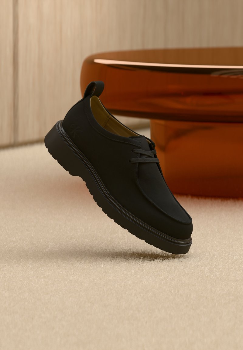 Chaussure décontractée en cuir noir avec lacets, semelle épaisse et languette de tirage, suspendue en l'air au-dessus d'un tapis beige avec un meuble arrondi marron en arrière-plan.