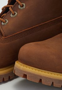 Brune læderstøvler med rund tå, metaløjet og brune snørebånd. Tekstureret overdel med syede detaljer og en tykk, rillet gummisål.