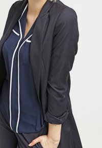 Marinblus med vit piping, lagd under en figurnära svart blazer. Blazern har en slank design och uppkavlade ärmar.