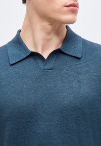 Homme portant un polo tricoté bleu foncé texturé, de profil, mettant l'accent sur le col et le haut du torse.