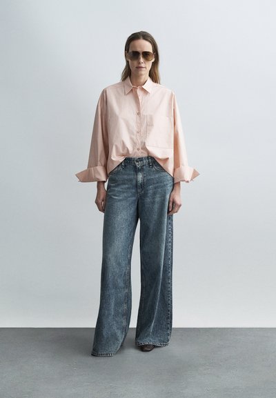 Hellrosa übergroßes Hemd mit Knöpfen und Tasche, kombiniert mit weit geschnittenen, ausgewaschenen blauen Jeans, abgerundet durch große braune Sonnenbrillen.