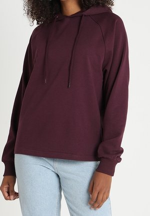 Frau trägt einen locker sitzenden, dunkelvioletten Hoodie mit Kordelzug und hellblaue Jeans, steht vor einem einfarbigen Hintergrund.