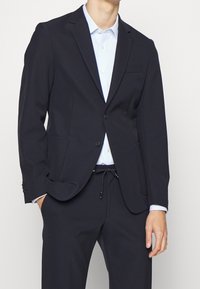 Mörkblå kavaj i mjukt tyg, med en bröstficka och två framfickor. Åtföljd av en ljusblå skjorta med krage.