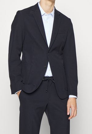 Giacca in blu scuro realizzata in tessuto liscio, con una tasca sul petto e due tasche frontali. Abbinata a una camicia azzurra con colletto.