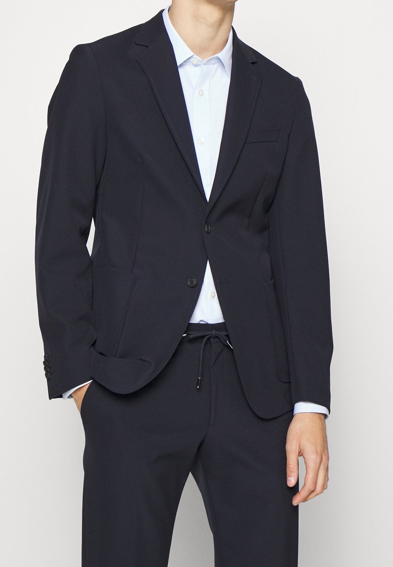 Mörkblå kavaj i mjukt tyg, med en bröstficka och två framfickor. Åtföljd av en ljusblå skjorta med krage.