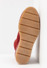 Red suede ankle boot with a textured beige rubber sole. The sole features horizontal grooves for grip and a circular logo imprint.