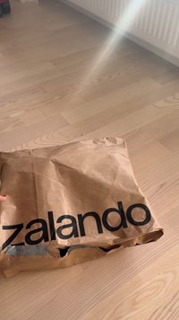 Saco de entrega Zalando de papel castanho amassado repousando no chão de madeira clara perto de um radiador branco.