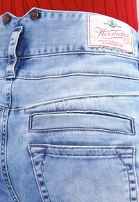 Světle modré džíny z denimu s texturovaným povrchem, s hnědou koženou nášivkou a dvěma zadními kapsami s horizontálním prošíváním.