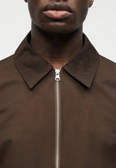 Κοντινό πλάνο του κάτω μέρους του προσώπου και του λαιμού ενός ατόμου που φοράει καφέ πουκάμισο με κεντρικό ασημένιο φερμουάρ και μυτερό γιακά.