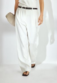 Bele, plisirane široke hlače iz gladkega materiala, v kombinaciji z črnim pasom. Oseba drži bež torbico in nosi ravne črne sandale.