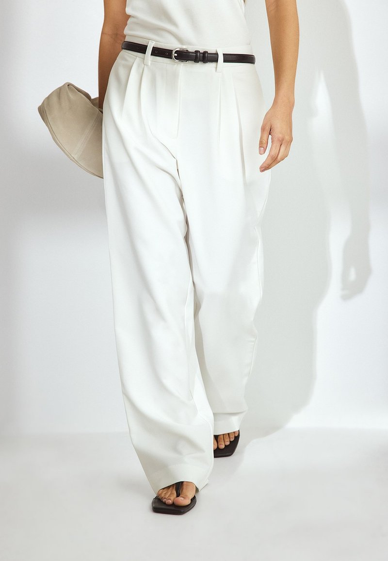 Bele, plisirane široke hlače iz gladkega materiala, v kombinaciji z črnim pasom. Oseba drži bež torbico in nosi ravne črne sandale.