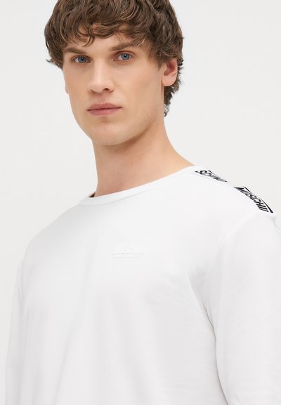 Valge pikkade varrukatega särk, millel on rinnal reljeefne logo ja mustad detailid õla ääres. Pehme puuvillane kangas, ümara kaeluse ja mugava lõikega.