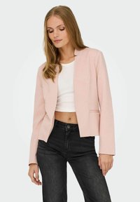 Femme portant un blazer rose pâle sur un haut blanc court et un jean noir taille haute, debout devant un fond uni.