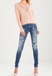 Maglione lavorato a maglia rosa chiaro con un motivo testurizzato, abbinato a jeans skinny blu consumati con strappi, e décolleté beige.