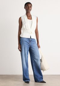 White sleeveless vest with a knot detail at the waist, paired with light blue wide-leg jeans and black pointed-toe shoes.