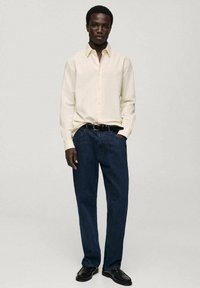 Light yellow, long-sleeve button-up shirt paired with dark blue denim jeans. Black belt and shoes complete the look. Simple, structured fit.