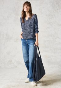 Blusa a fantasia navy con scollatura a V e maniche arrotolate, abbinata a jeans a gamba larga di un blu chiaro e sneakers bianche. Borsa tote nera.
