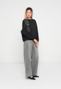 Kvinne med langt hår iført en svart oversized hettegenser med hvit linjekunst, grå vid bukse og svarte spisse heels, står på hvit gulv.