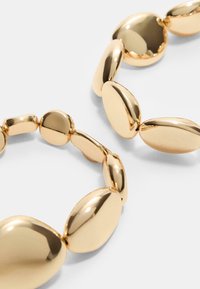 Bracciale tono oro con anelli ovali irregolari, dotati di una superficie liscia e lucida, che riflette la luce e mette in risalto un design senza soluzione di continuità.