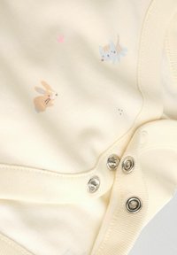 Kremowy body dla niemowląt z zatrzaskami, z małymi nadrukami zajączka i kotka w delikatnych pastelowych kolorach.