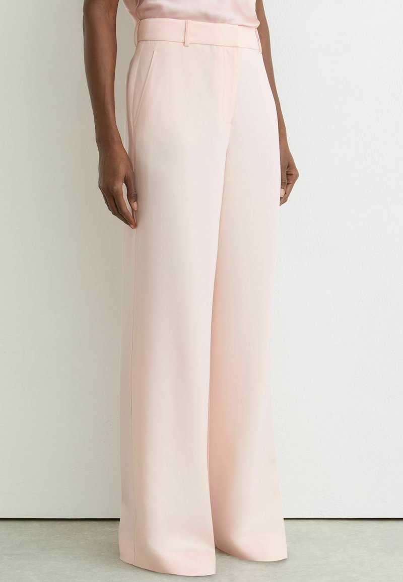 Nő világos rózsaszín, magas derekú, bőszárú nadrágban, aki fehér fal és szürke padló előtt áll.