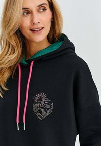 Černá mikina s kapucí, zeleným podšíváním a růžovými šňůrkami. Výrazný design srdce se slunečními a vlnovými grafikami v pastelových barvách.