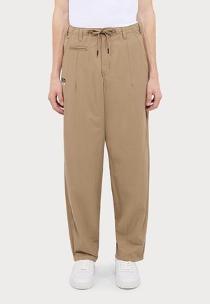 Beige afslappede bukser med snøre og plisseringer, en lille forlomme, sidestykke-detalje, båret med hvide sneakers og en hvid top.
