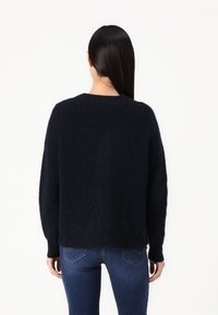 Kvinna med långt svart hår som bär en mörkblå fluffig tröja och blå jeans, stående vänd bort från kameran mot en enkel vit bakgrund.