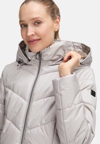 Ljusgrå, vadderad jacka med hög krage, dragkedjestängning och texturerad foder. Funktioner inkluderar chevronstygn och en logotyp patch på ärmen.