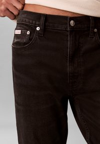 Gros plan d'une personne portant un jean en denim marron foncé avec une étiquette Calvin Klein visible près de la poche et un haut blanc côtelé.