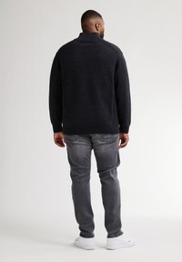 Dunkelgrauer Strickpullover mit hohem Kragen, geripptem Saum und Schulterdetails, kombiniert mit grauen Jeans und weißen Turnschuhen.