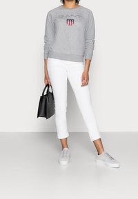 Sudadera gris con logo y escudo bordados, pantalones blancos con puños, zapatillas deportivas grises y un bolso negro. Diseño de atuendo simple y casual.