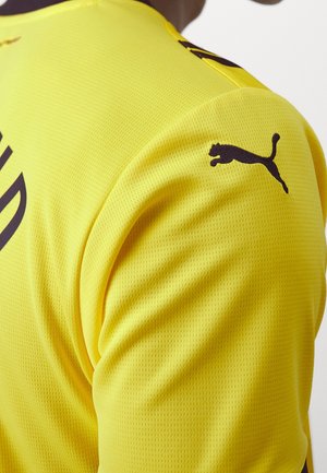 Nahaufnahme der Schulter eines gelben Sportshirts mit einem schwarzen Puma-Logo und einem teilweisen Text auf dem Stoff.